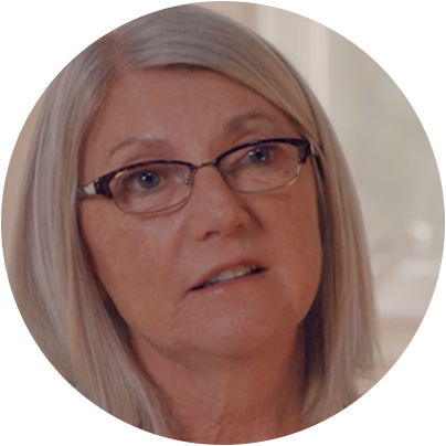 Pictured is a circular headshot of Marie. She is around 50 years old. She has black glasses on and medium length grey hair. There is a grey background behind her.