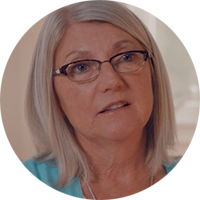 Pictured is a circular headshot of Marie. She is around 50 years old. She has black glasses on and medium length grey hair. She is wearing a blue shirt and there is a grey background.