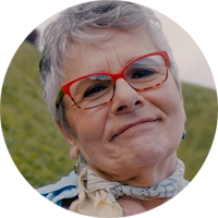 Pictured is a circular headshot of Marilyn. She is about 55-65 years old. She has short grey hair, red glasses, and a yellow and blue scarf tied around her neck. In the background is a grassy field. 